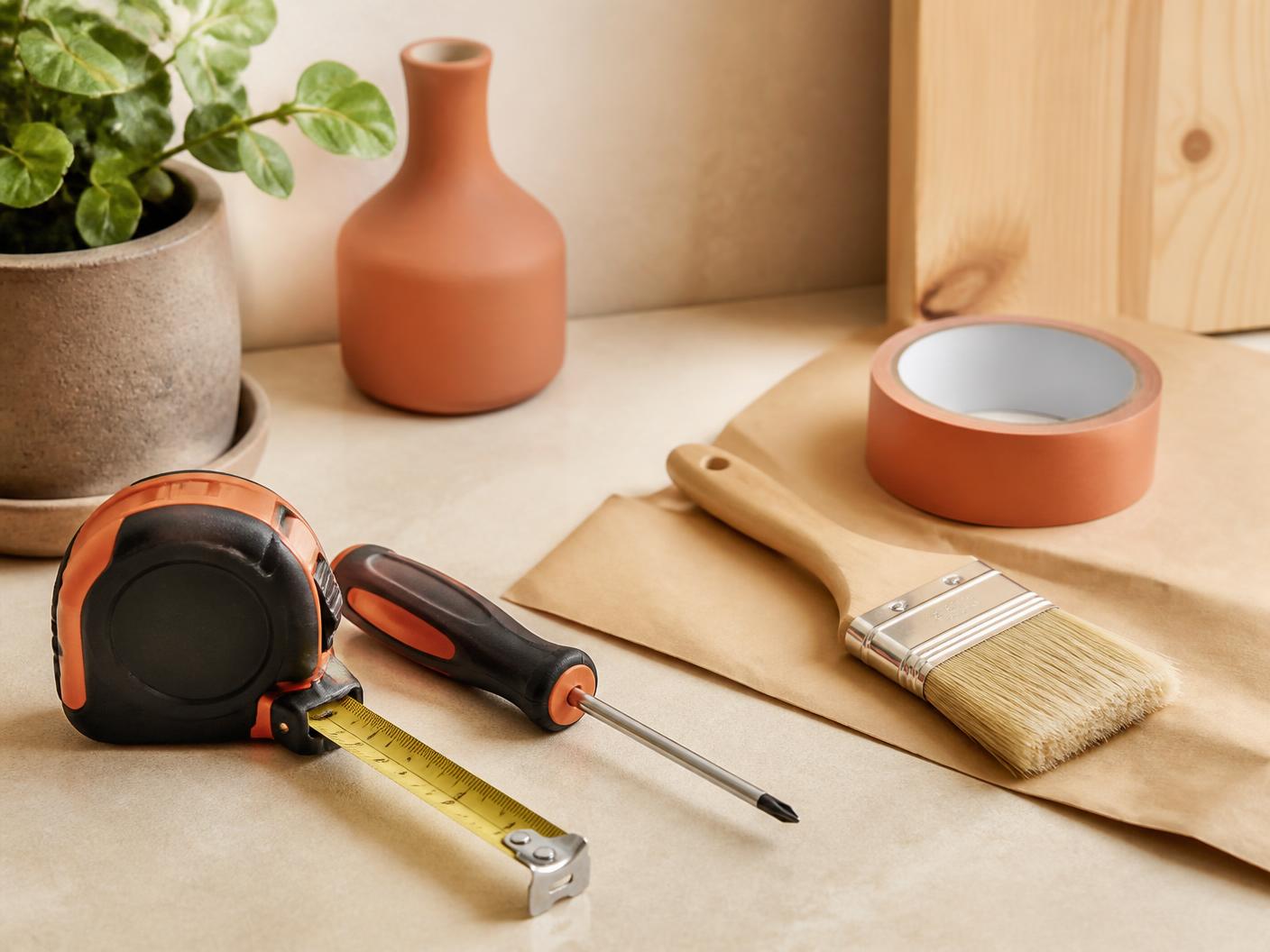 Tools & Home Improvement category editorial photograph — a measuring tape, screwdriver, paint brush and roll of tape on a work surface. Lifestyle imagery representing the category, not a photograph of the listed product.